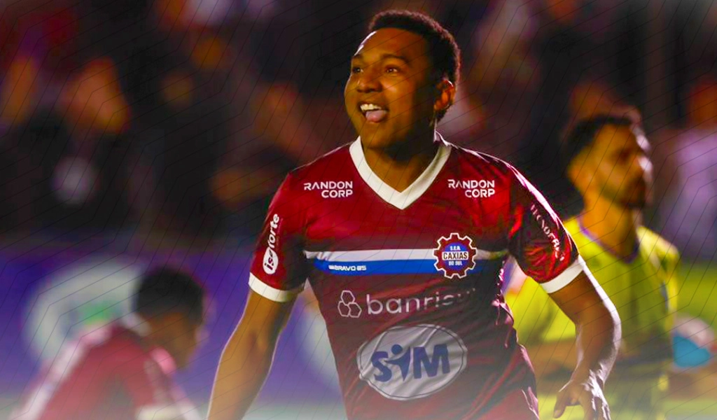 Jogador de futebol com camisa do SER Caxias correndo com sorriso no rosto - ao fundo outro jogador e o rbitro da partida.